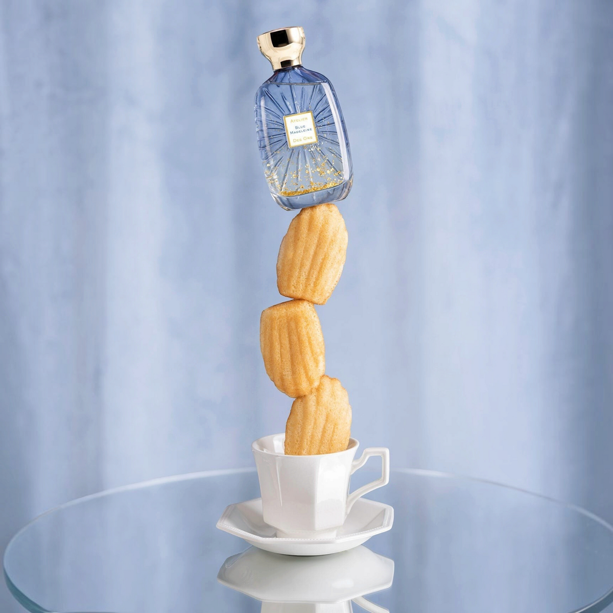 Shop Rescue Spa Blue Madeleine perfume bottle balanced atop a stack of madeleine cakes in a teacup on a glass table.