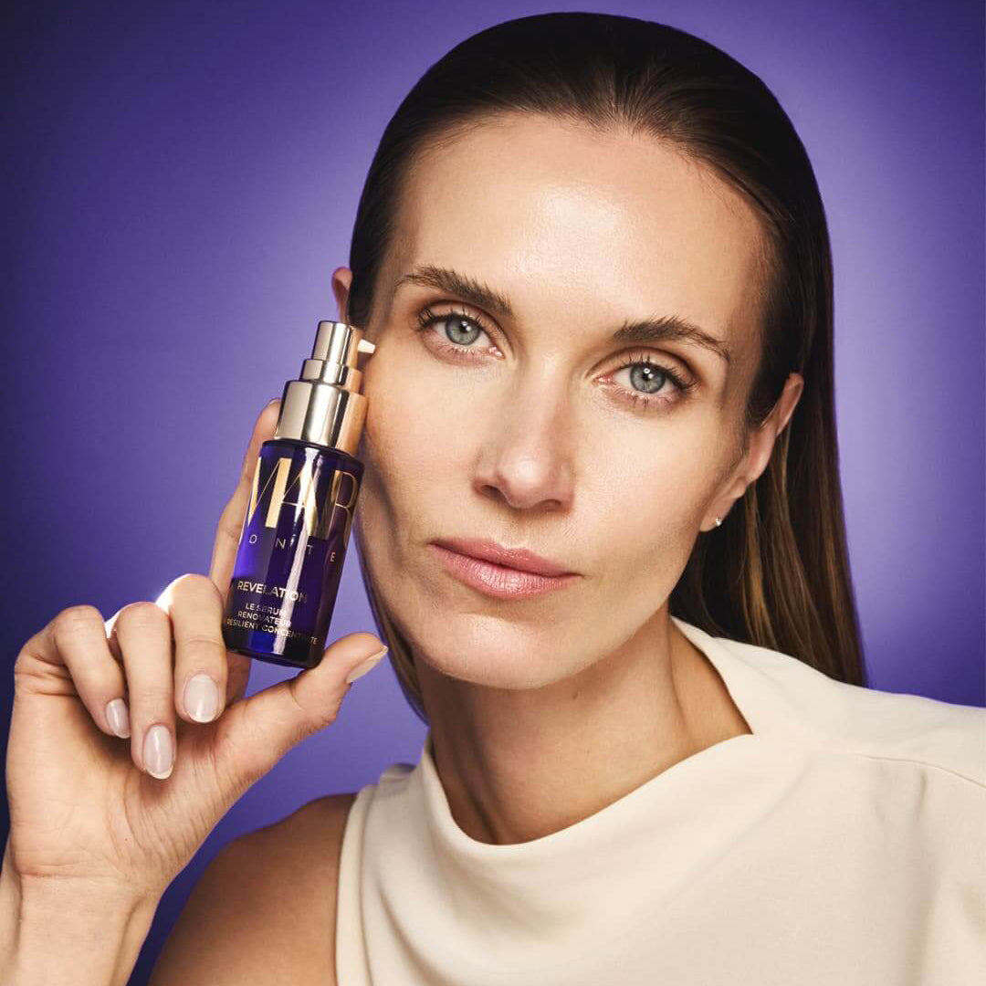 A woman with clean, bare skin holds a small purple cosmetic bottle with a gold pump against a solid purple background. The bottle has gold lettering that appears to read &