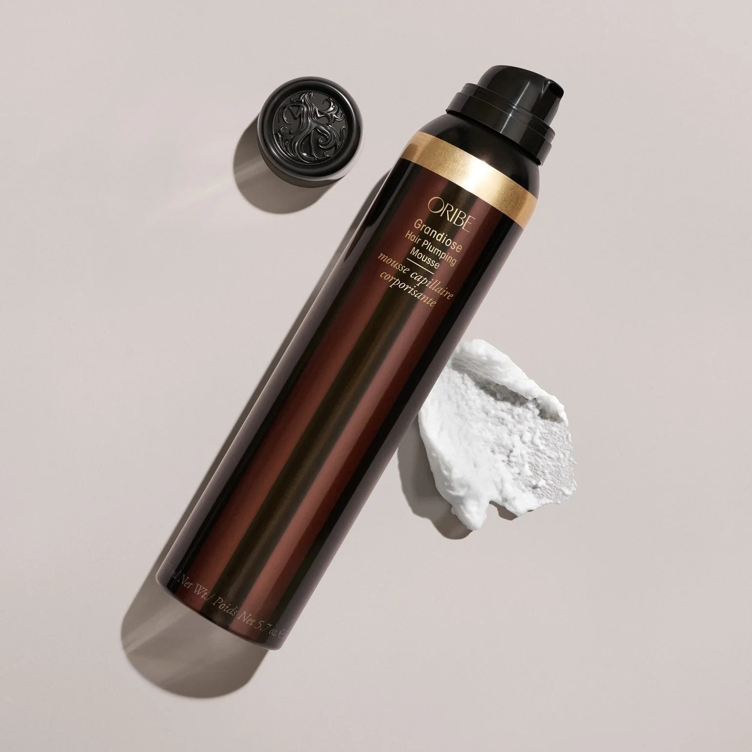 Shop Rescue Spa Oribe Grandiose Hair Plumping Mousse bottle with a dollop of white foam on a neutral background.