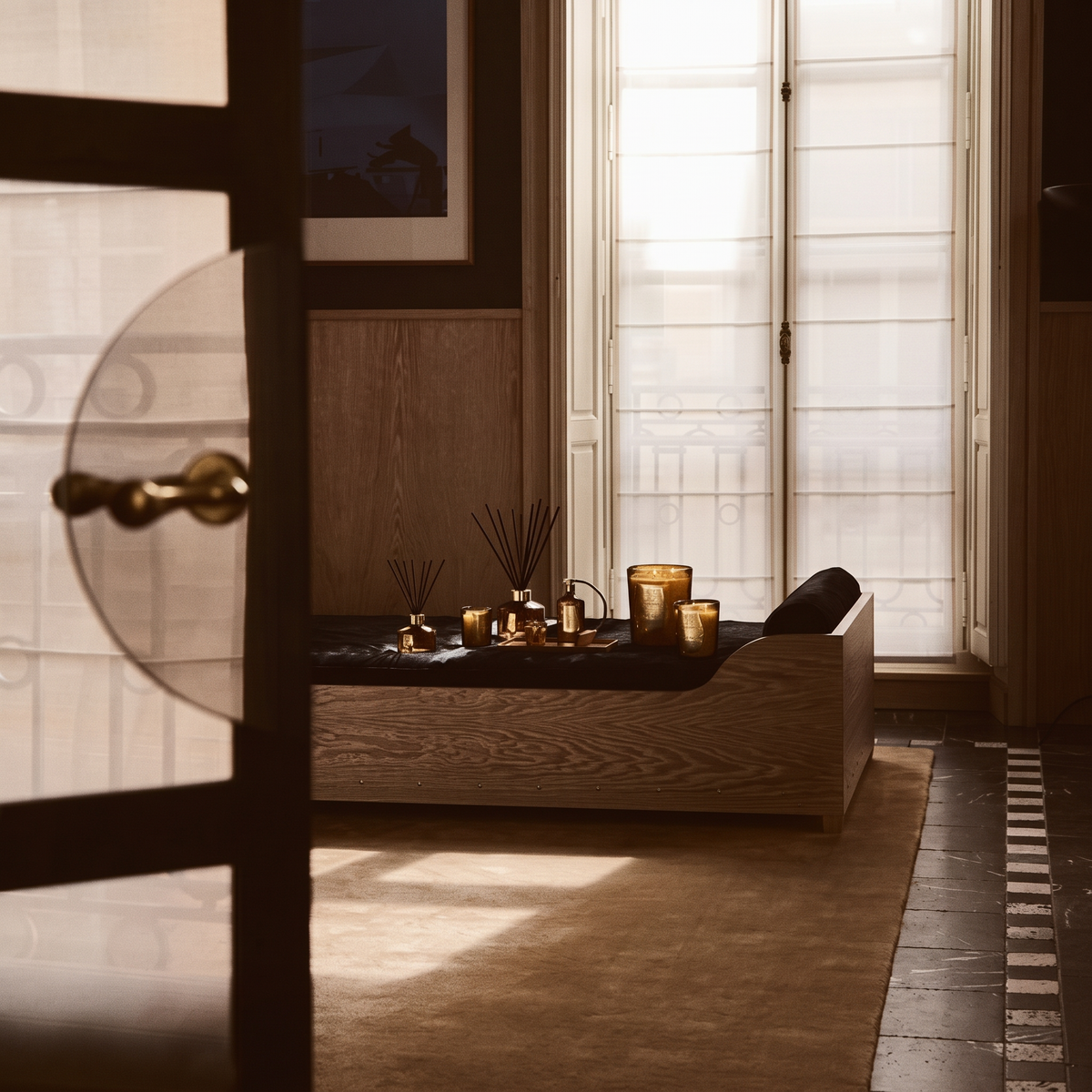 A styled interior scene with a low wooden bench or daybed against a sunlit window, decorated with gold-toned candle holders and reed diffusers. A dark cushion rests on the seat, and warm natural light streams in from the right side.