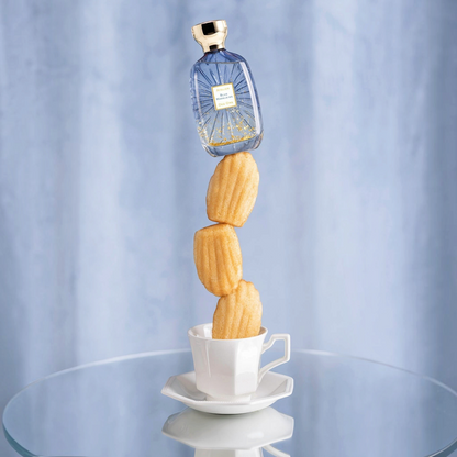 Shop Rescue Spa Blue Madeleine perfume bottle balanced atop a stack of madeleine cakes in a teacup on a glass table.