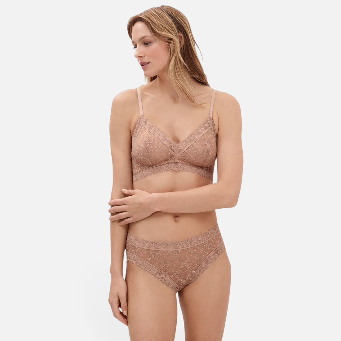 A woman modeling a matching lace bra and panty lingerie set in a dusty rose color, photographed against a solid light gray background.