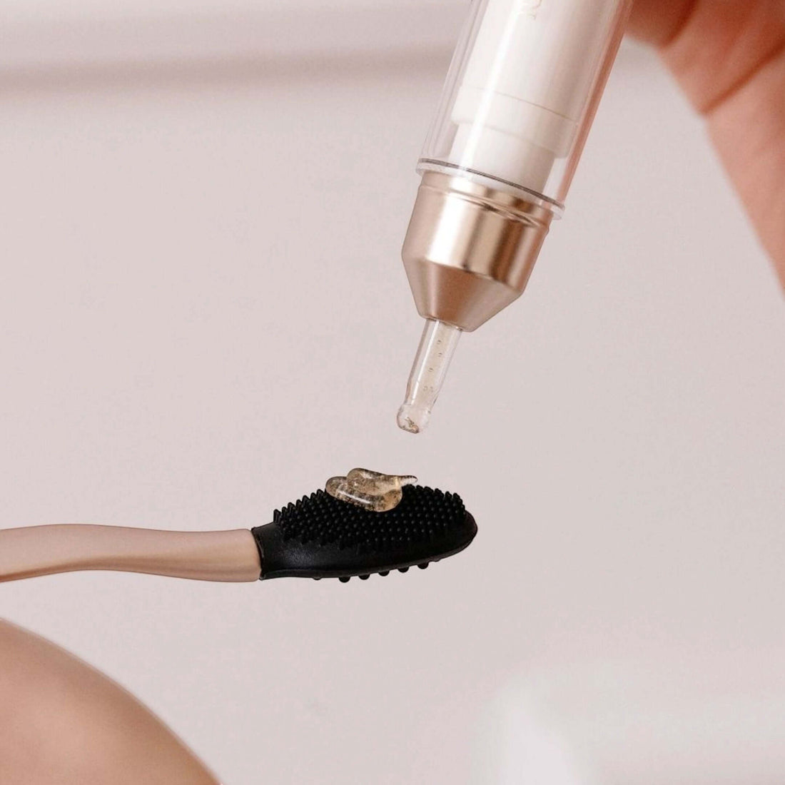 A close-up of a hand holding a clear dropper above a black silicone scrubber brush with a beige handle, dispensing a clear gel or liquid onto the brush head.