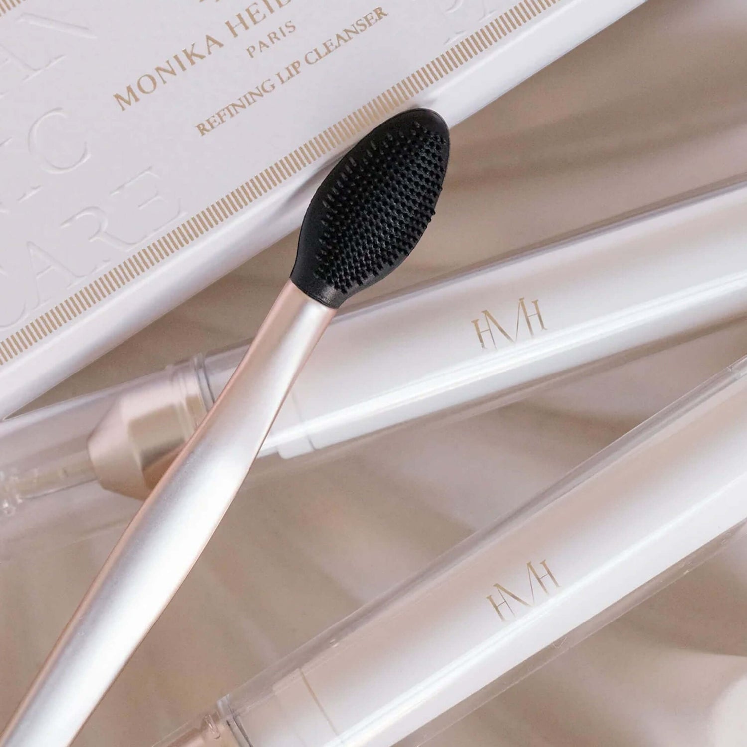 A close-up of a lip cleansing brush with a black bristle head and pale pink handle, resting on a white box labeled Monika Heide Paris Refining Lip Cleanser. Two white cosmetic tubes with gold &