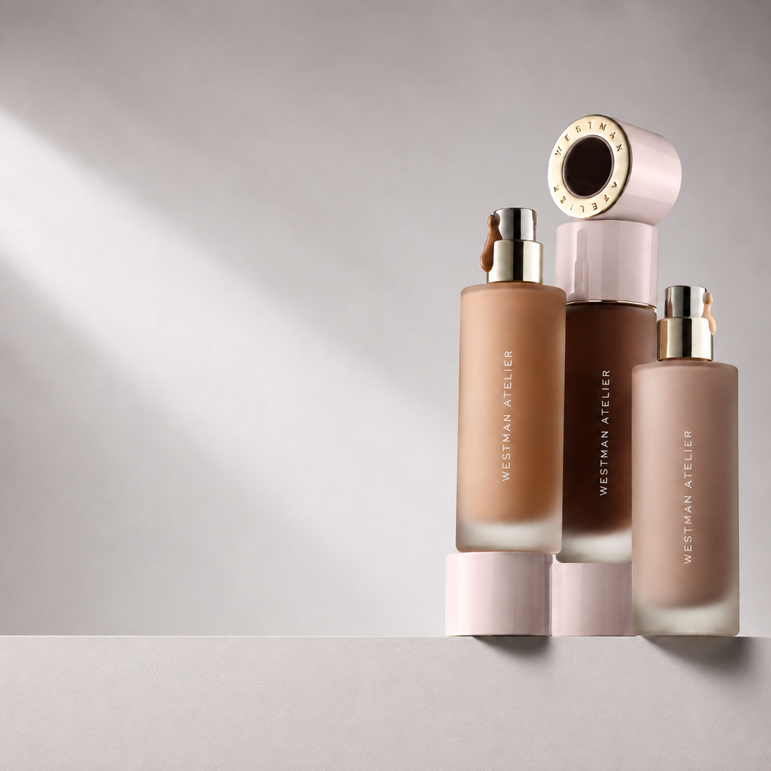 Three Westman Atelier cosmetic bottles in frosted glass with gold caps, arranged on a light gray surface against a matching gray background with soft diagonal light.