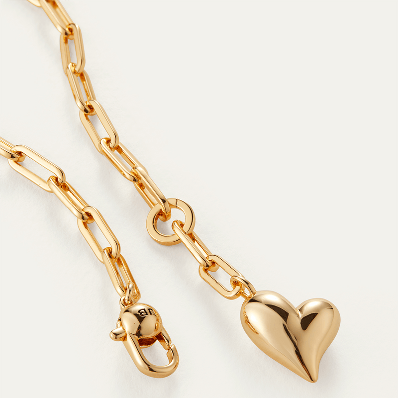 A close-up of a gold chain necklace featuring a large paperclip link chain and a polished heart-shaped pendant, shown on a light off-white background.
