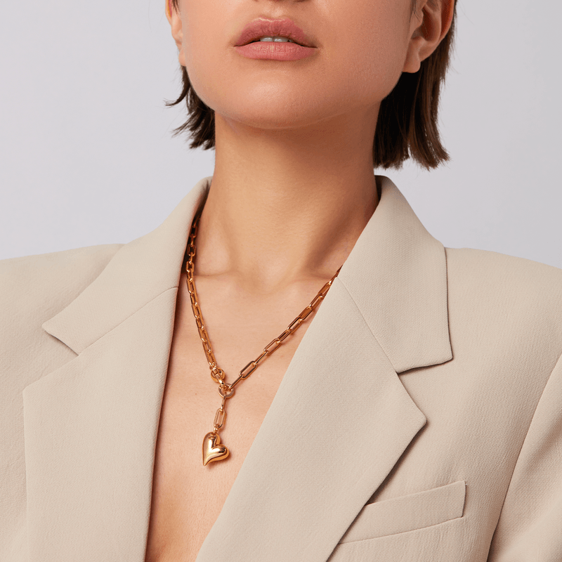 A close-up of a model wearing a gold heart pendant necklace with a chunky chain, styled with a beige blazer.