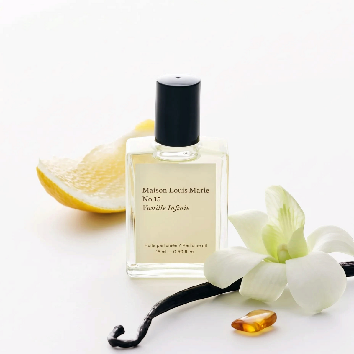 A bottle of Maison Louis Marie No.15 Vanille Infinie perfume oil, styled with a lemon slice, a vanilla flower, a vanilla bean, and a small amber droplet on a clean white background.
