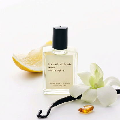 A bottle of Maison Louis Marie No.15 Vanille Infinie perfume oil, styled with a lemon slice, a vanilla flower, a vanilla bean, and a small amber droplet on a clean white background.