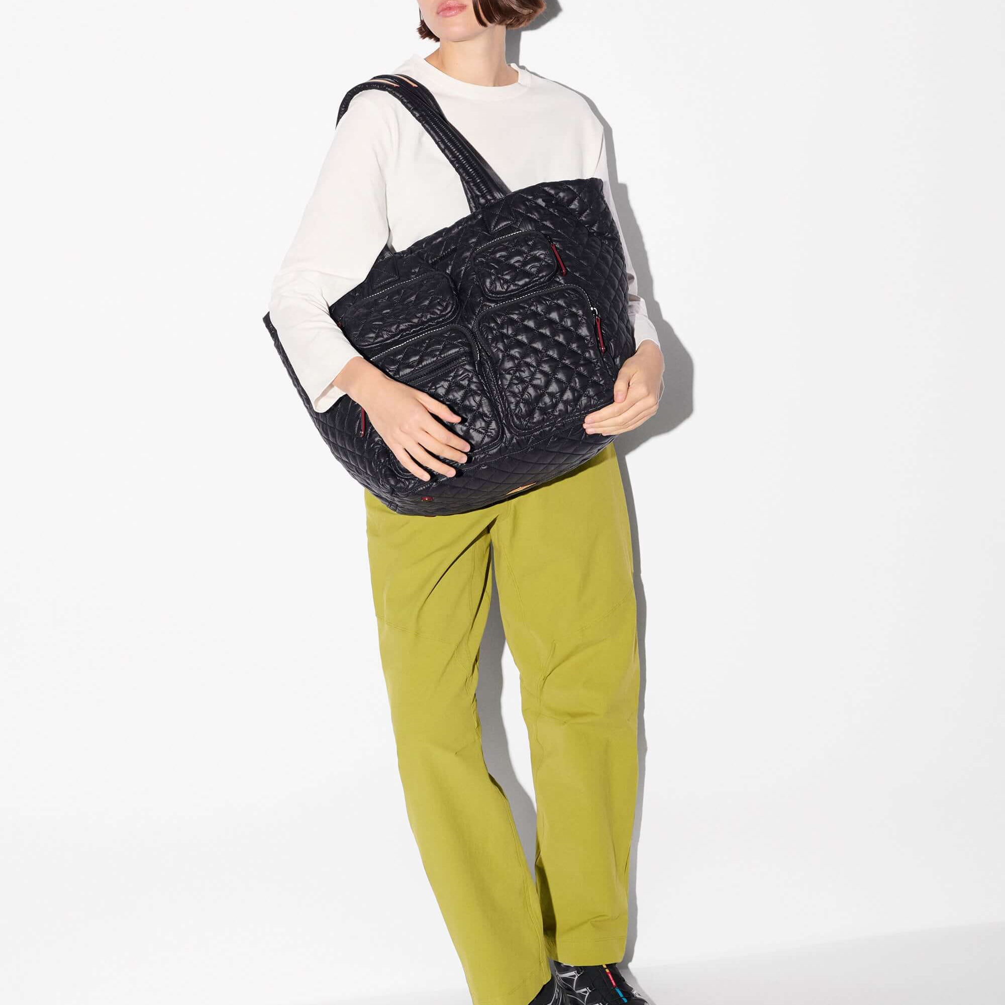 A model wearing a white long-sleeve shirt, olive green pants, and black sneakers carries a large black quilted shoulder bag with multiple front pockets against a plain white background.