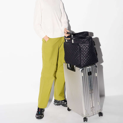 A person wearing a white long-sleeve top and olive green pants stands beside a silver rolling suitcase, holding a black quilted travel bag. The scene is photographed against a clean white studio background.