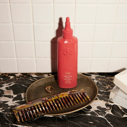 Oribe Bright Blonde Radiance &amp; Repair Treatment bottle with a tortoiseshell comb and gold ring on a marble surface.