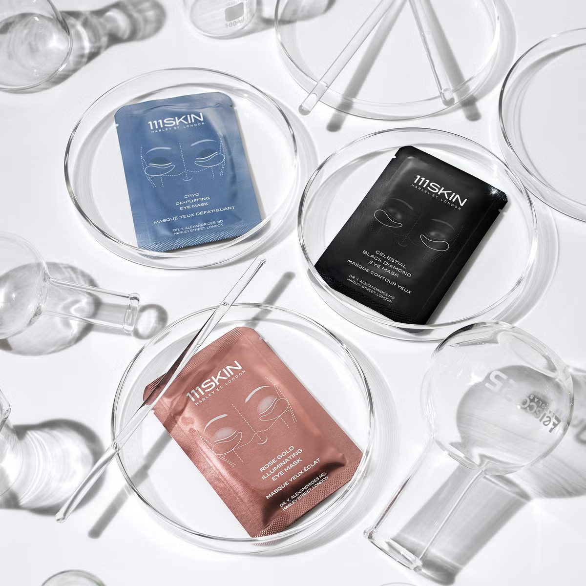 Shop Rescue Spa 111SKIN hydrogel eye masks in rose gold, blue, and black packaging arranged in glass laboratory dishes.