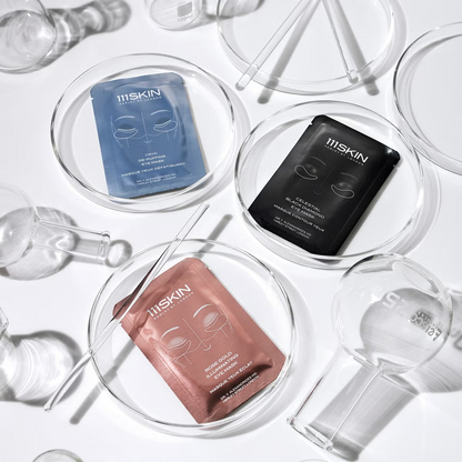 Shop Rescue Spa 111SKIN hydrogel eye masks in rose gold, blue, and black packaging arranged in glass laboratory dishes.