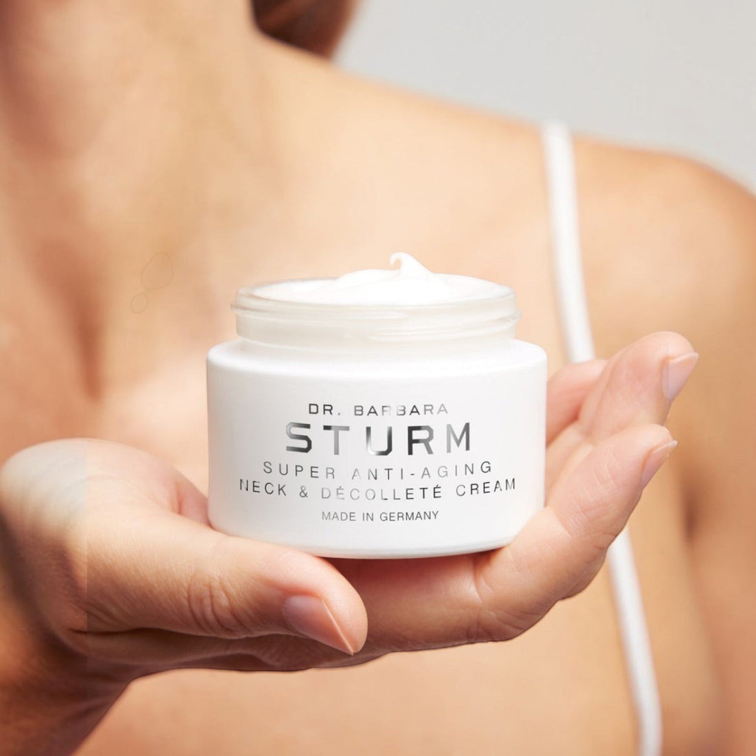 Shop Rescue Spa Dr. Barbara Sturm Super Anti-Aging Neck &amp; Décolleté Cream in a white jar held in a hand