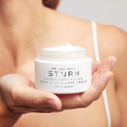 Shop Rescue Spa Dr. Barbara Sturm Super Anti-Aging Neck &amp; Décolleté Cream in a white jar held in a hand
