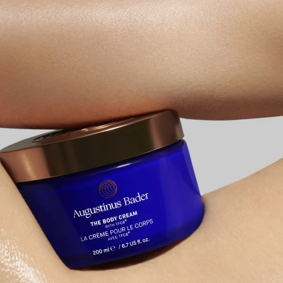 Shop Rescue Spa Augustinus Bader The Body Cream in a blue jar held against hydrated, glowing skin.