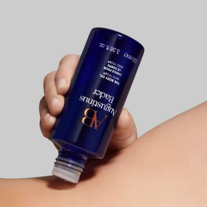 Shop Rescue Spa Augustinus Bader The Body Oil in a blue bottle being applied to hydrated, glowing skin.