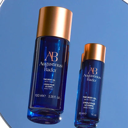 Shop Rescue Spa Augustinus Bader The Body Oil bottles in blue glass with metallic gold caps on a reflective surface.