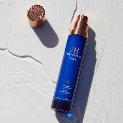 Shop Rescue Spa Augustinus Bader The Cream Cleansing Gel in a blue bottle with a copper pump on a wet surface.