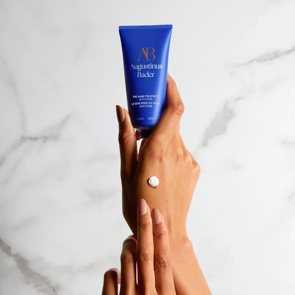 Shop Rescue Spa Augustinus Bader The Hand Treatment in a blue tube held by hands with a dab of white cream