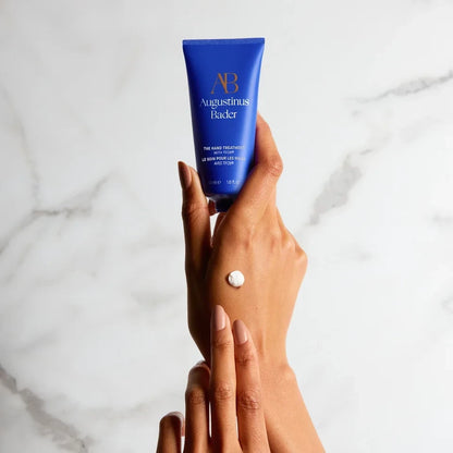 Shop Rescue Spa Augustinus Bader The Hand Treatment in a blue tube held by hands with a dab of white cream
