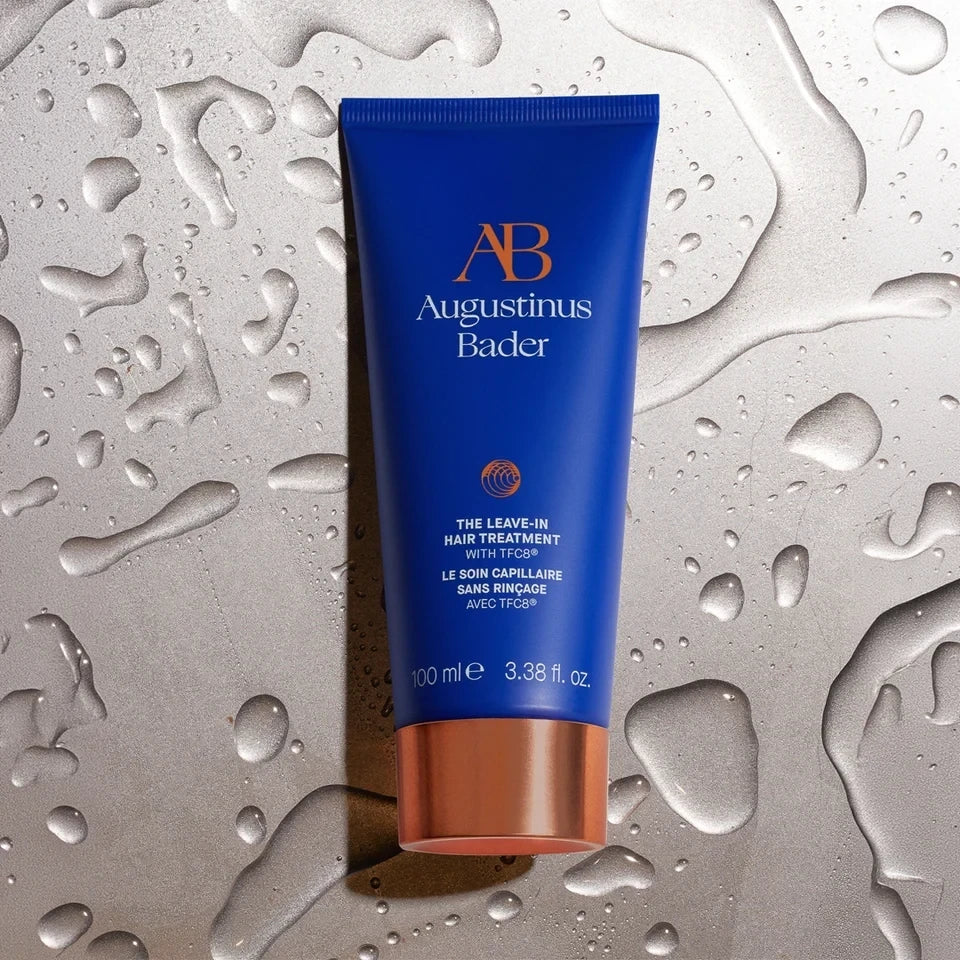 Shop Rescue Spa Augustinus Bader The Leave-In Hair Treatment in a blue tube on a metallic surface with water droplets.