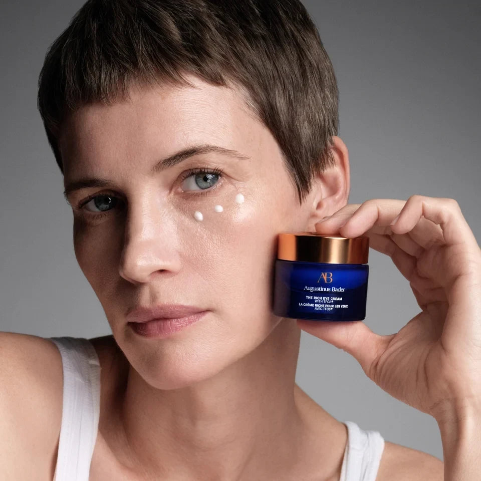 Shop Rescue Spa model holding Augustinus Bader The Rich Eye Cream jar with dots of cream applied under her eye
