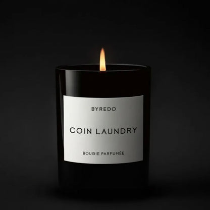 Shop Rescue Spa Byredo Coin Laundry candle in a black glass jar with a lit wick. Floral scent with notes of aldehyde, pink pepper, rose, neroli, peony, violet, woods, musk, and sandalwood.