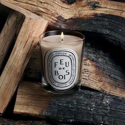 Shop Rescue Spa Diptyque Feu de Bois candle in a glass vessel, lit and placed against a background of charred logs.