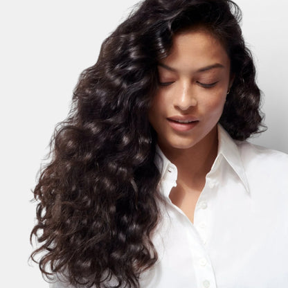 Shop Rescue Spa Gold Lust Nourishing Hair Oil on dark wavy hair of a woman with a white shirt.