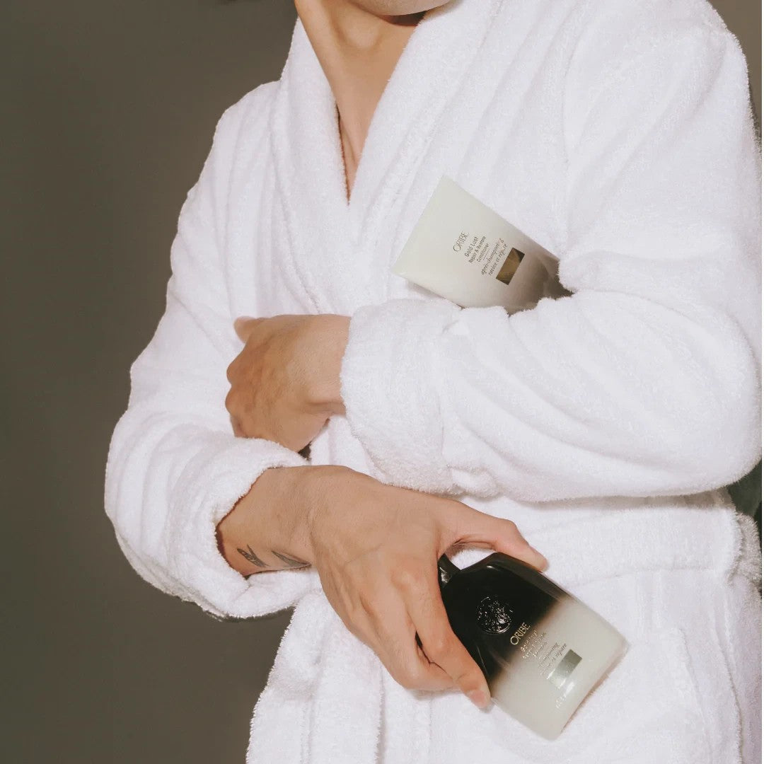 Shop Rescue Spa Oribe Gold Lust shampoo and conditioner bottles held by a person wearing a white plush bathrobe.