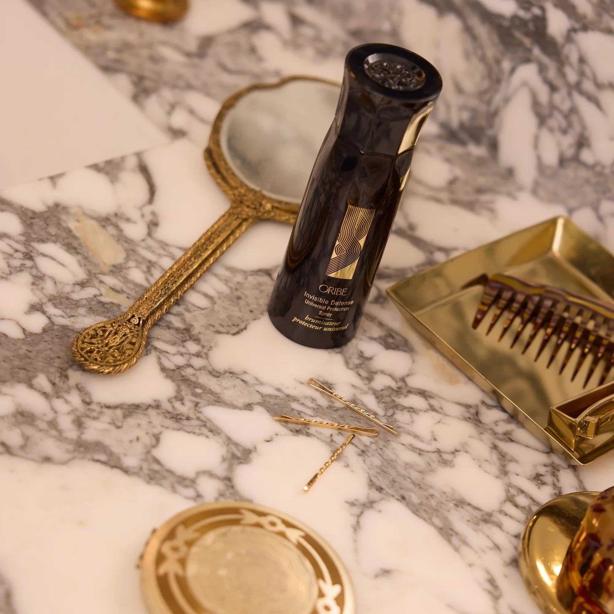 Shop Rescue Spa Oribe Invisible Defense hair protection spray bottle on a marble vanity with gold accessories.