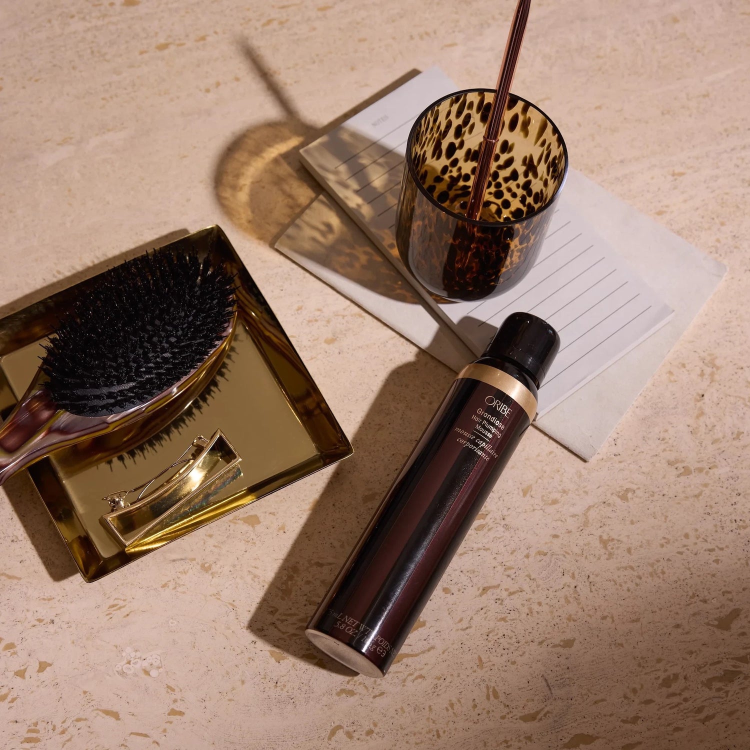 Shop Rescue Spa Oribe Grandiose Hair Plumping Mousse bottle on a vanity with a hairbrush and leopard print glass.