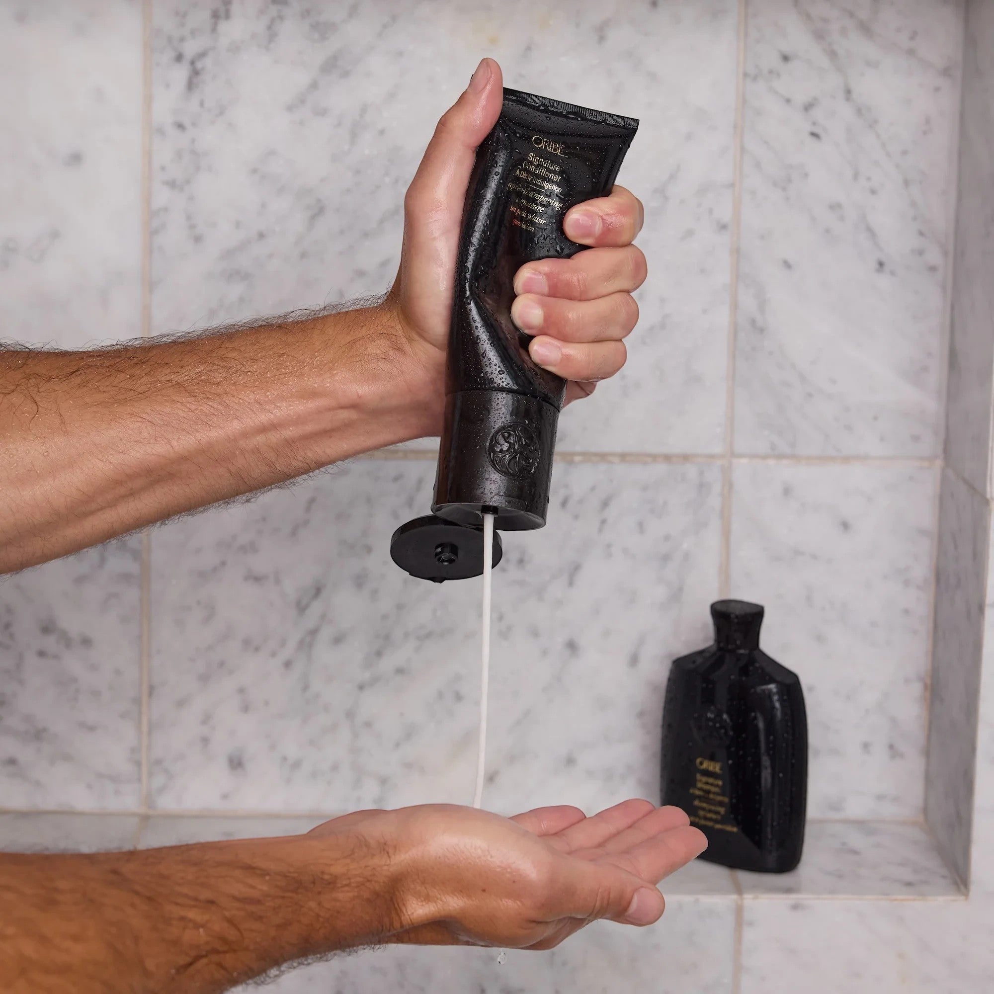Shop Rescue Spa Oribe Signature Conditioner being squeezed from a black tube into a hand in a marble shower