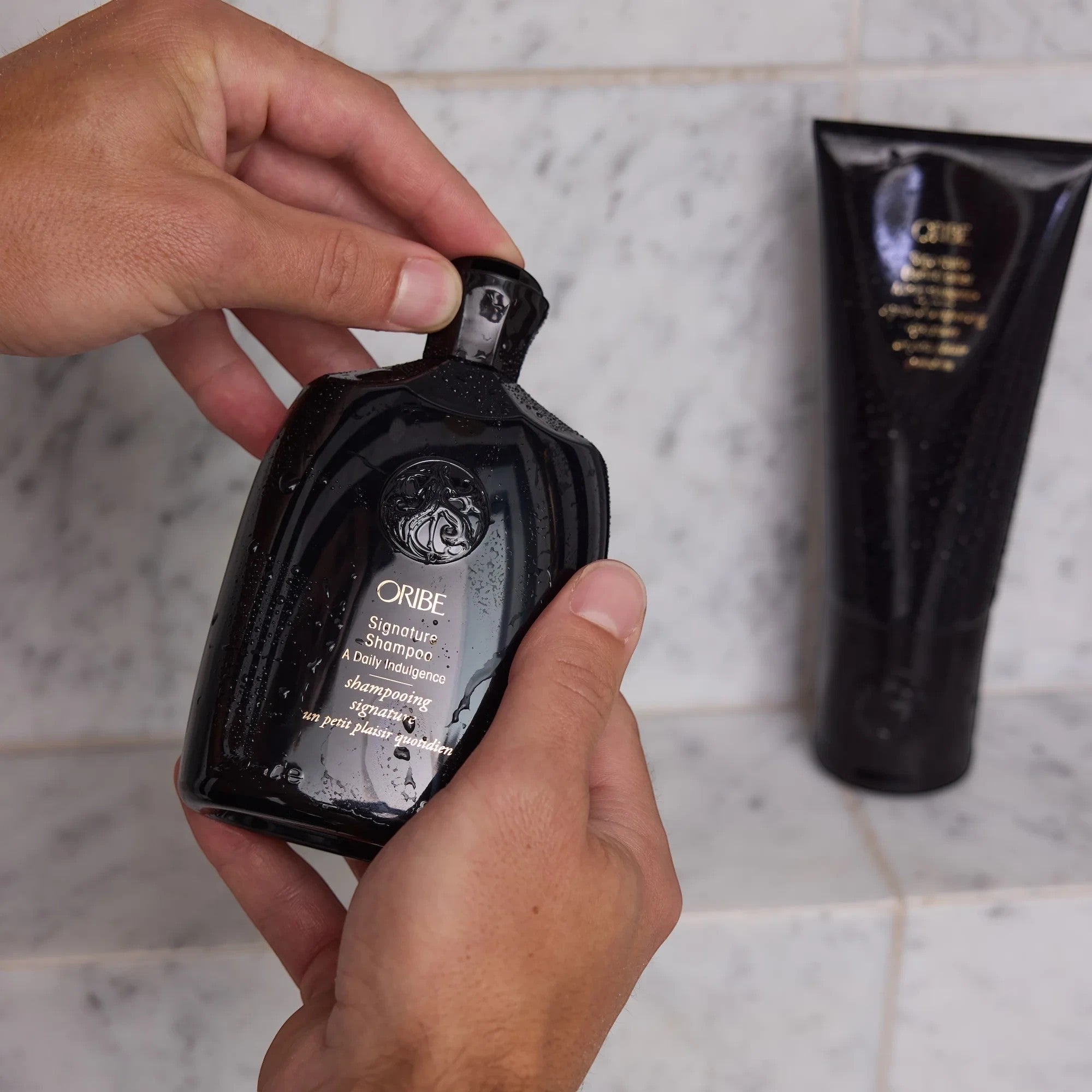 Shop Rescue Spa Oribe Signature Shampoo bottle held in hands in a marble shower with water droplets on the surface.