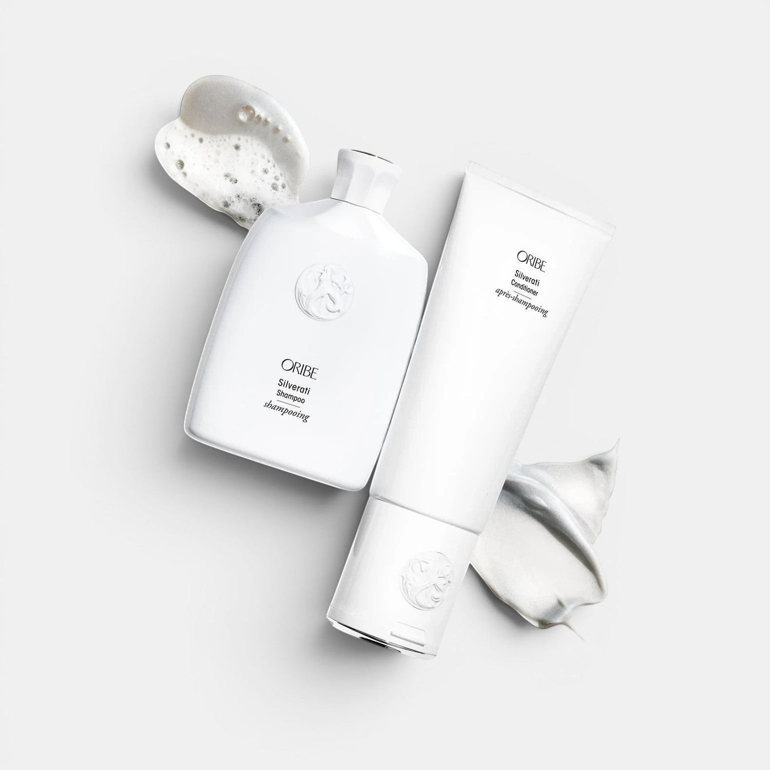 Oribe Silverati Shampoo and Conditioner bottles with white foam and cream on a white background.