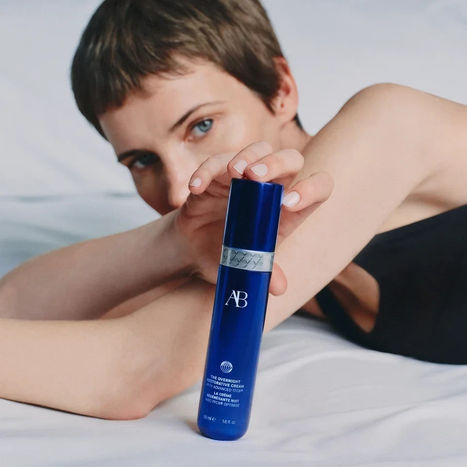 Shop Rescue Spa The Overnight Restorative Cream in a blue bottle held by a woman resting on white bedding.