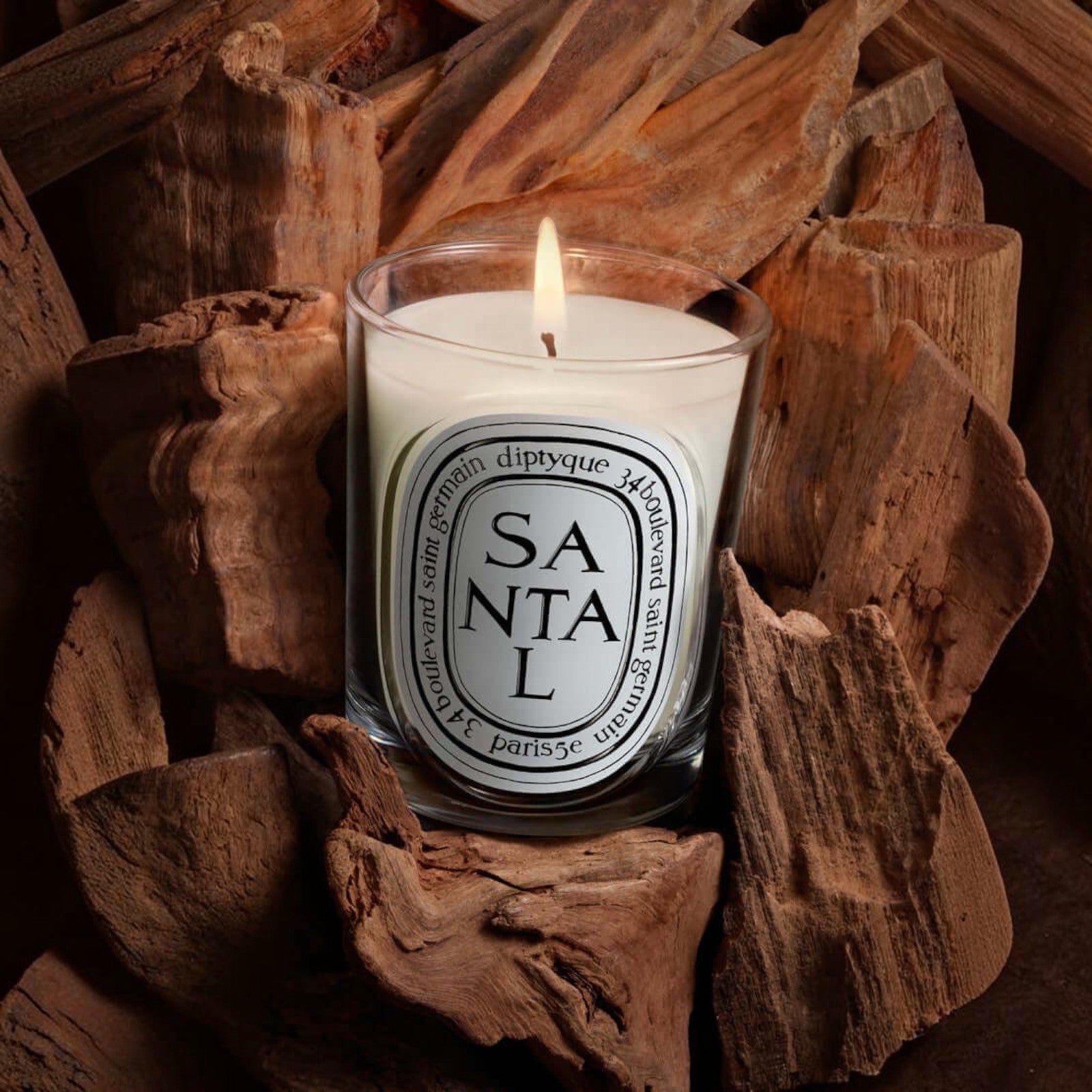 Shop Rescue Spa Diptyque Santal scented candle in a glass jar, surrounded by raw sandalwood pieces.