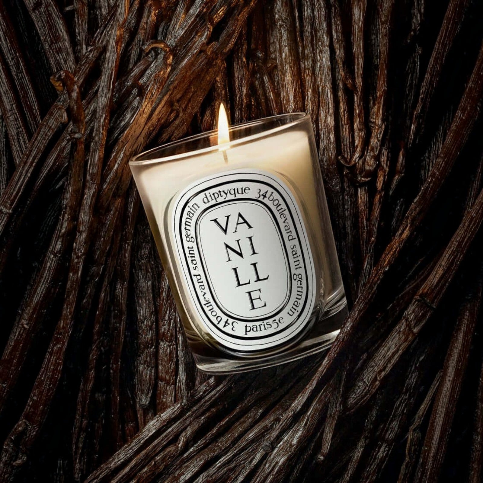 Shop Rescue Spa Diptyque Vanille scented candle in a glass vessel, glowing with a flame against dark vanilla pods.