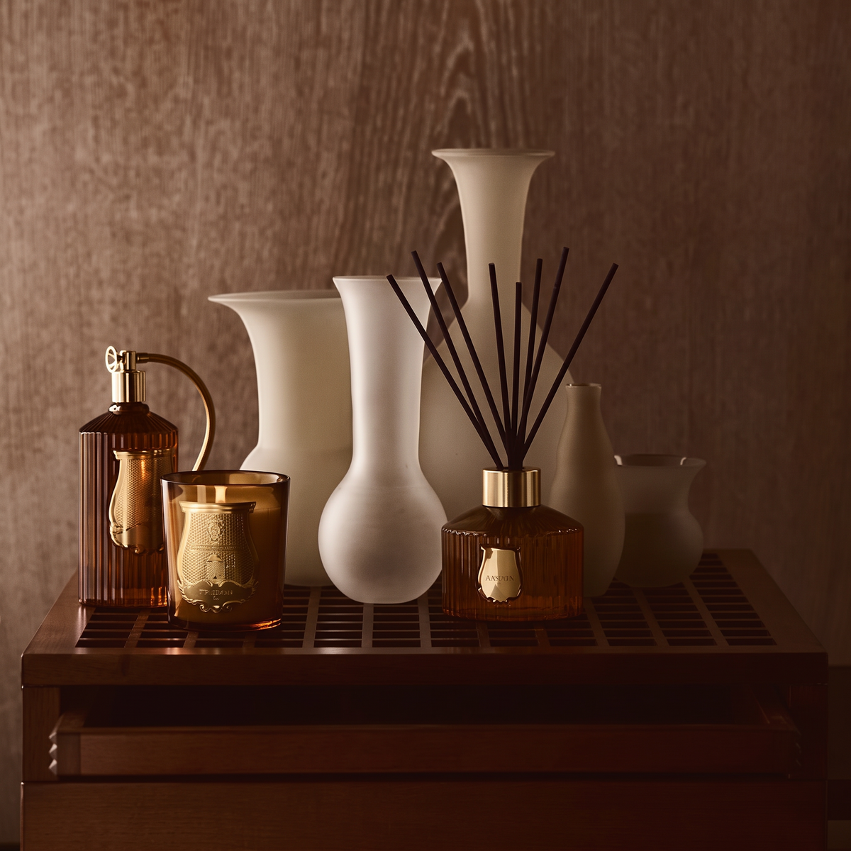 A styled collection of home decor items on a dark wooden tray, including matte white ceramic vases, amber glass bottles, and a reed diffuser, set against a warm brown wood-paneled background.