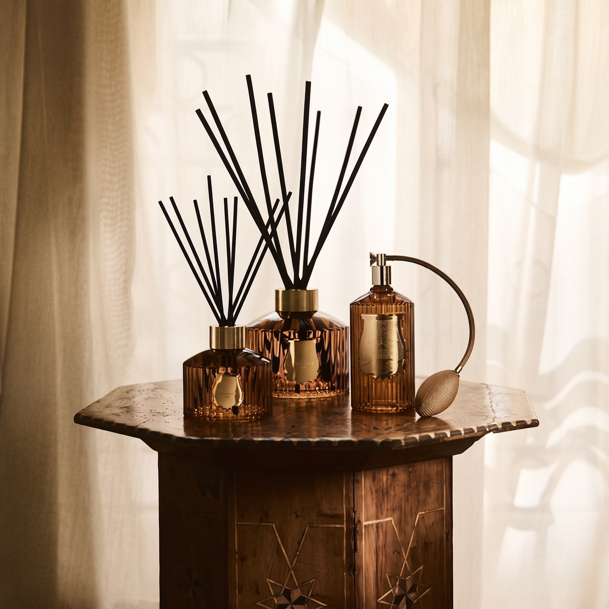 Three amber glass reed diffusers with black reeds and a matching amber atomizer bottle arranged on a rustic wooden table in front of soft cream curtains.