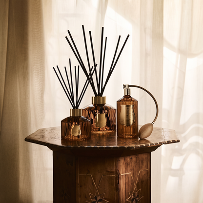 Three amber glass reed diffusers with black reeds and a matching amber atomizer bottle arranged on a rustic wooden table in front of soft cream curtains.