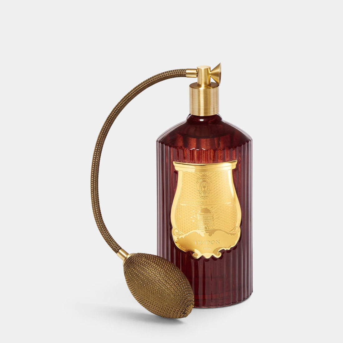 A vintage-style perfume bottle with a ribbed dark red glass body, a large gold crest on the front, and a brass pump and flexible bulb nozzle, photographed on a light gray background.
